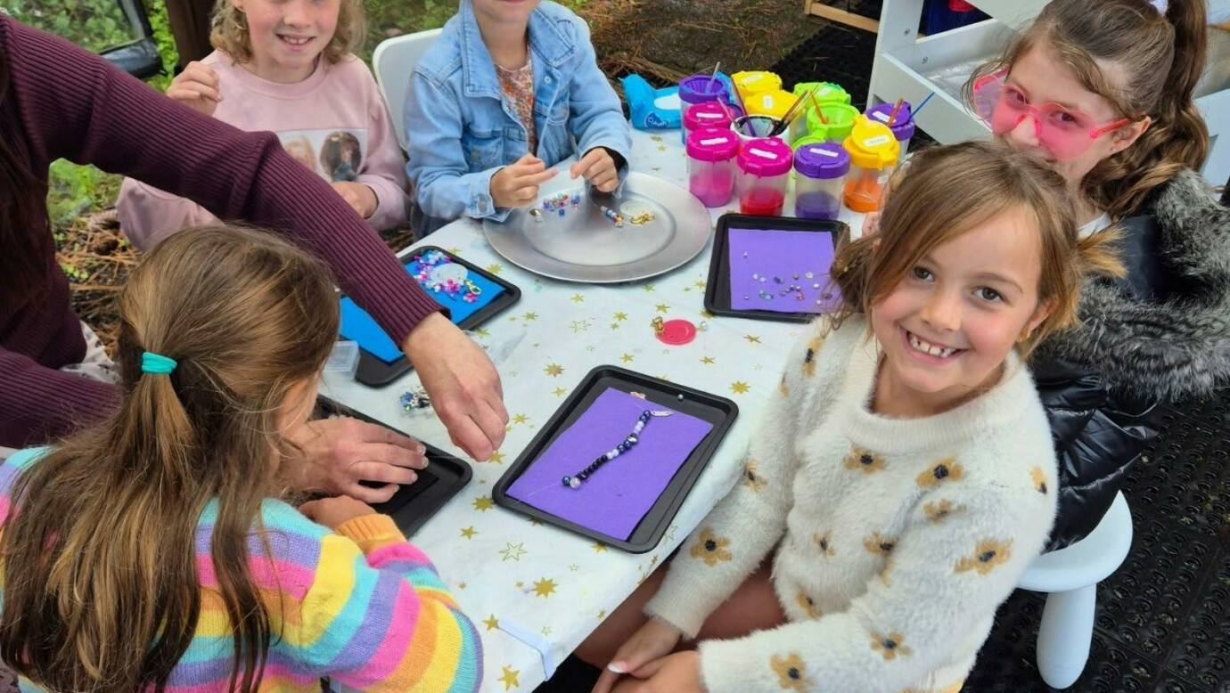 A group of young girls sitting around a table outdoors making beaded bracelets, with colourful mater