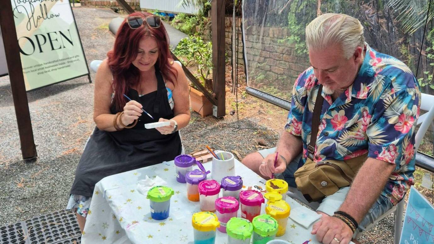 A man and woman sitting at a painting table with bright paint jars, both focused