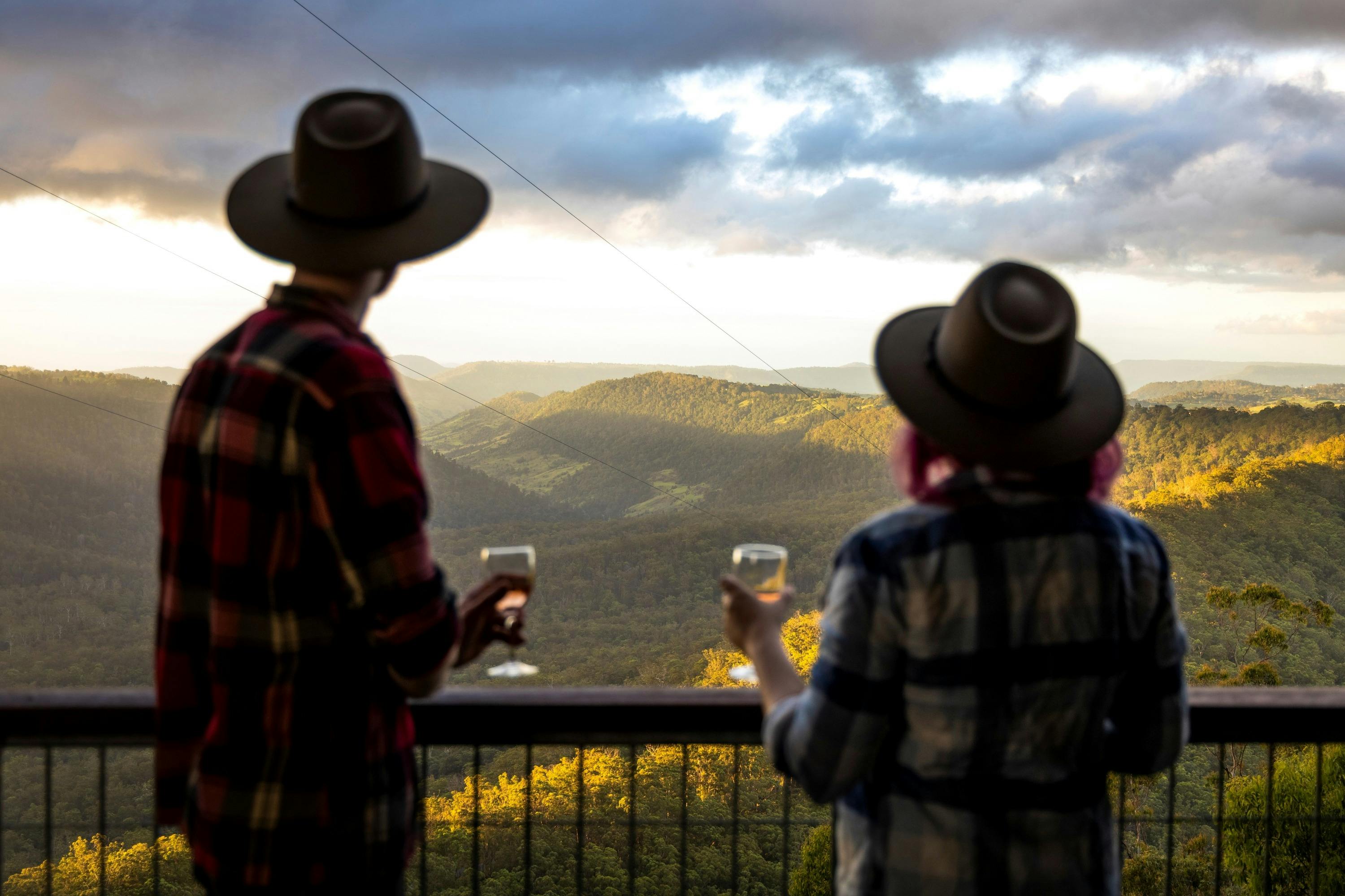 A couple stand at a balcony railing with a glass of wine in their hand looking at the valley below