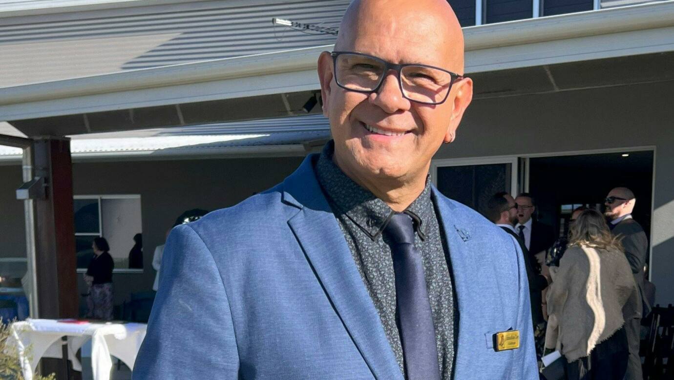 A man in a blue suit and tie stands outdoors, smiling warmly in front of a modern building during a