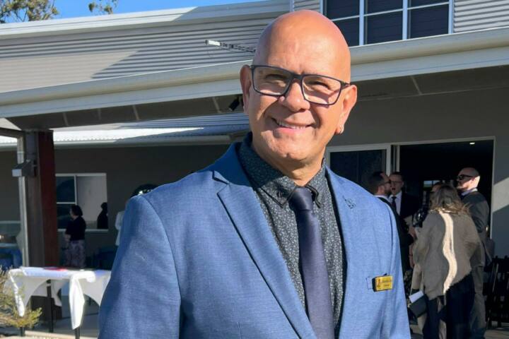 A man in a blue suit and tie stands outdoors, smiling warmly in front of a modern building during a
